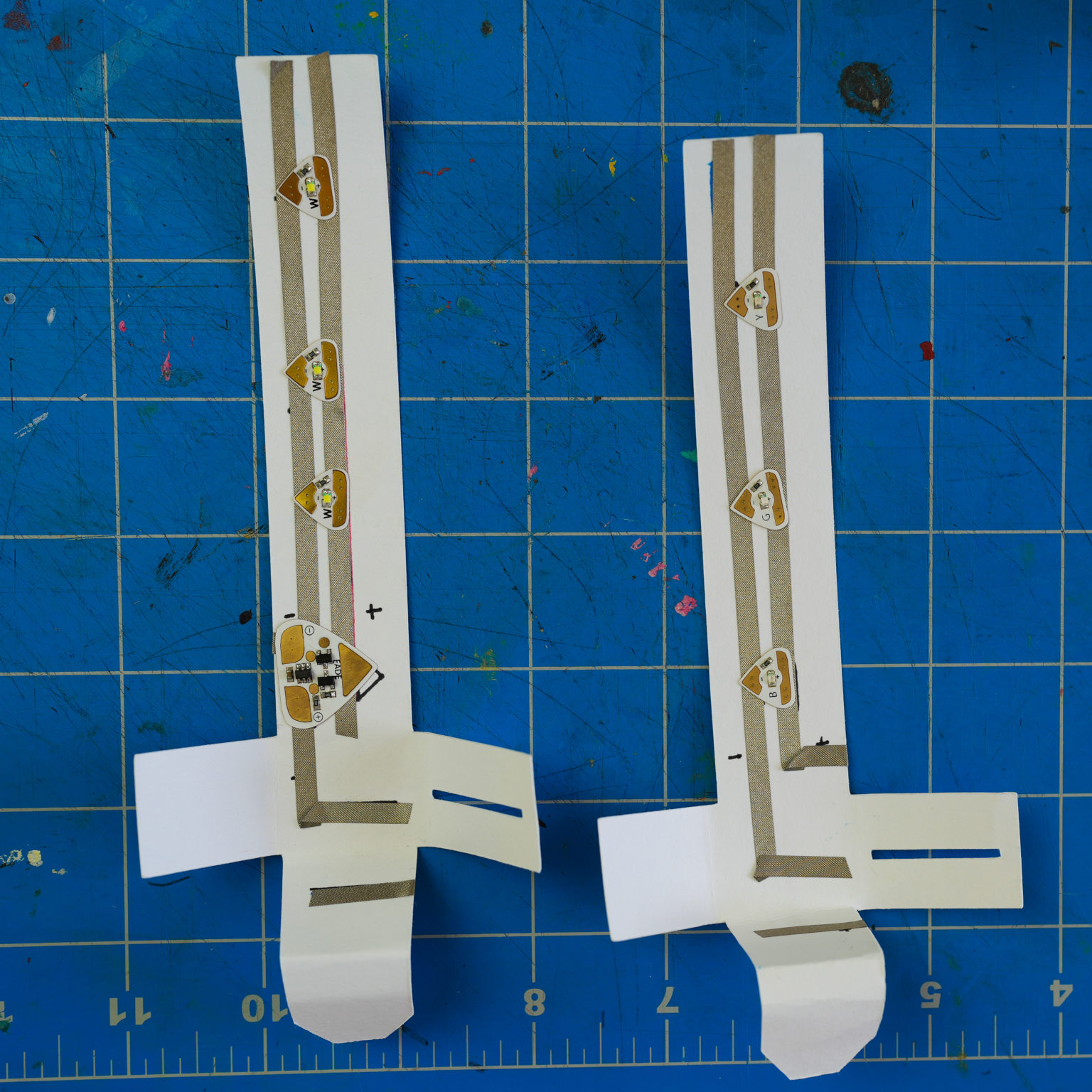 Two foldable light strips with three tabs extending from the bottom, rest upon a blue craft mat. The one of the left contains 3 Circuit Sticker LEDs and an Add-on Effects Sticker. The one of the right contains 3 LEDs. No battery has been placed in the holders.