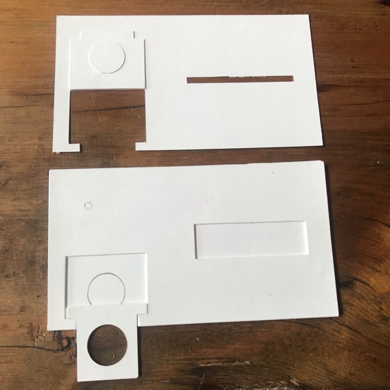Two plain, white parts of a card rest upon a wooden table. The top piece has a flap that is designed to rest upon the top of a coin cell battery. The bottom piece contains a tray made of paper with a hole large enough for a con battery to sit in.