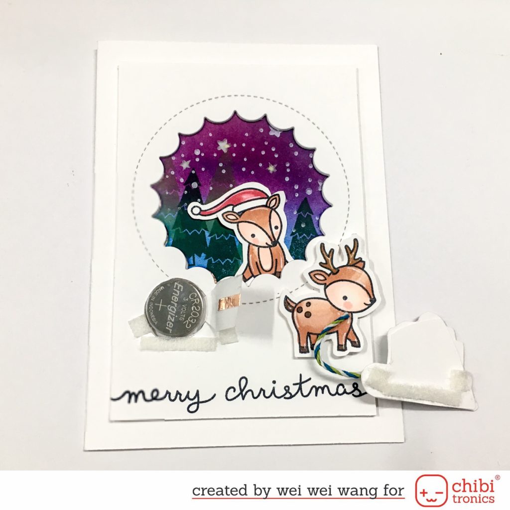 A white card with a purple medallion in the middle sits upon a white surface. In the center of the medallion is a squirrel wearing a Santa hat. To the left of the squirrel is coin cell battery that is touching part of a circuit. A flap on the right would complete the circuit if it were pressed down upon the battery. The back side of a paper door, adhered with Velcro tape, rests on the right side of the card.