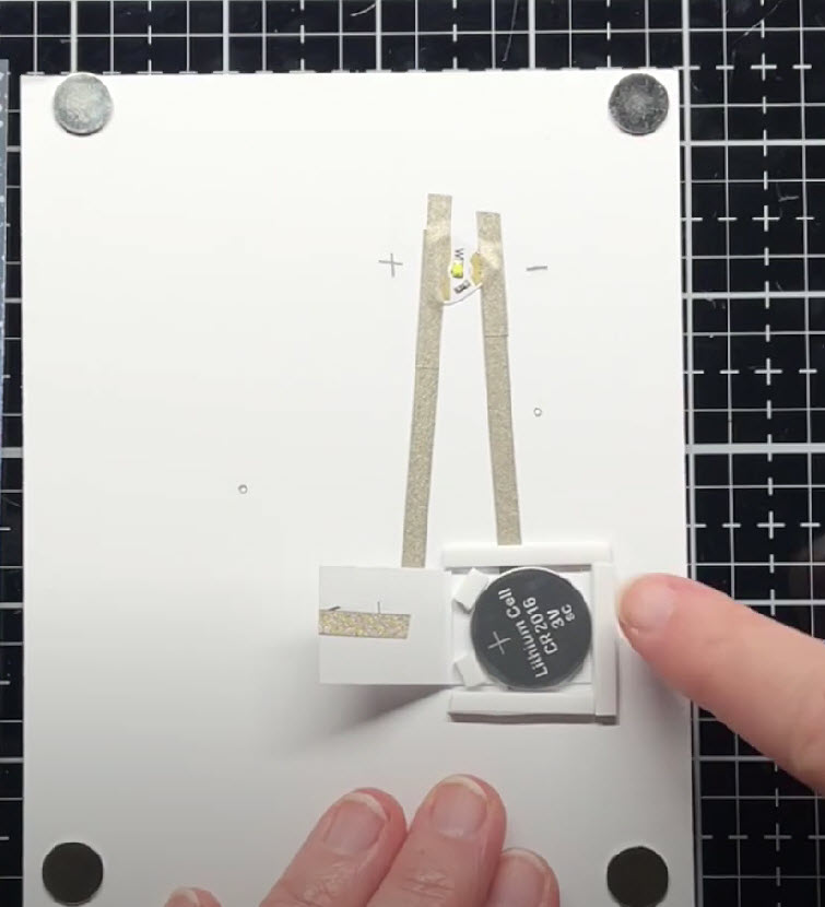 The inside of a white cardstock card panel has four round disk magnets attached in each corner. In the center of the card, a coin cell battery sits on top of a piece of conductive fabric tape that connects with the negative, pointy end of a Circuit Sticker LED. A paper flap to the right of the battery has another piece of conductive fabric tape wrapped around it and connecting to the broad, positive side of the LED. Three pieces of foam tape in the same of a three-sided square encase the battery.