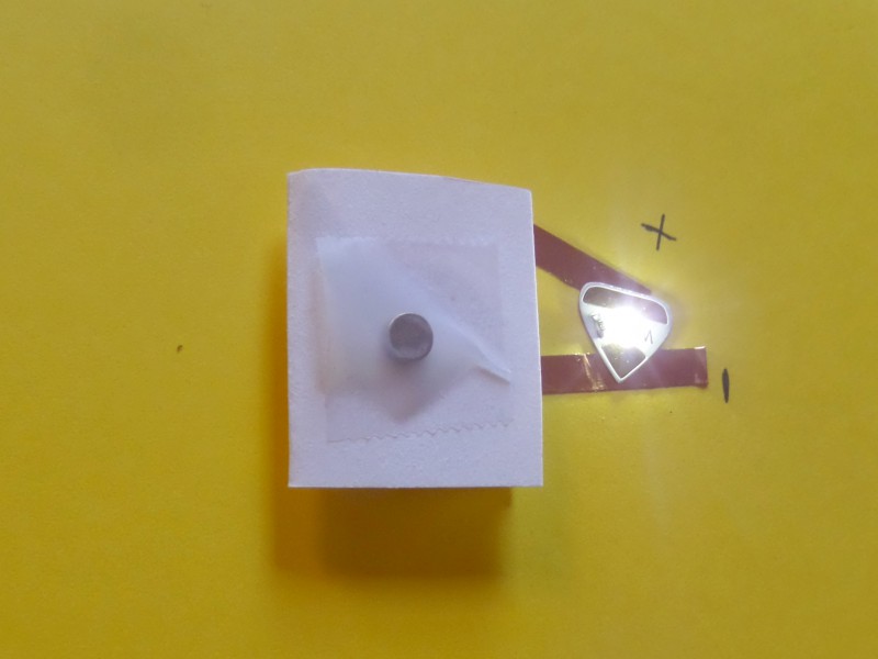 A folded piece of paper with a round disc magnet taped to the top with Scotch Tape connects to the positive and negative leads of a white Circuit Sticker LED, which is glowing.