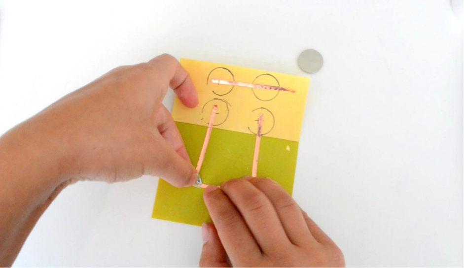 A yellow piece of paper has been labeled with four circular outlines of a coin cell battery. The outlines are placed in the shape of square, but they are not touching The top two outlines are connected together with a piece of copper tape. The one in the bottom left corner has been marked with minus sign and is connected to the negative end of a Circuit Sticker LED. The circular outline on the bottom right side of the paper has been labeled with a plus sing and connects to the positive end of the LED. A battery rests on the table in the right hand corner.