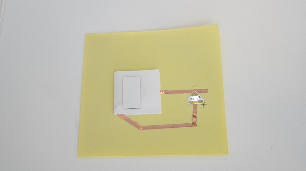 A folded piece of paper is attached to the top center of a yellow piece of paper. A rectangle of sheet magnet rests on top, suggesting the presence of a coin battery inside. To the right, a white Circuit Sticker LED glows. It connects to the hidden battery with copper tape.